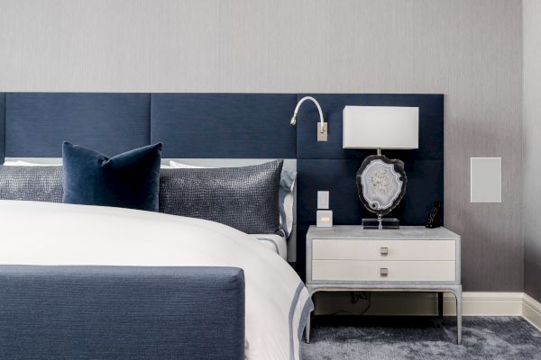 A modern bedroom with a blue and white color scheme featuring a bed, navy headboard, nightstand, lamp, and decorative items placed on the nightstand.