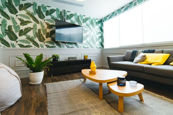 A modern living room with a gray sofa, yellow pillows, a TV on a wall with green leaf wallpaper, a plant, and wooden tables and flooring.