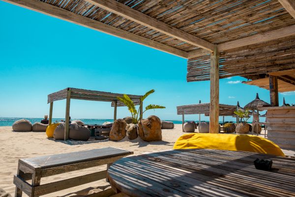 A sunny beach scene with shaded seating, umbrellas, and bean bags on the sand. The sky is clear and the ocean is visible in the background.