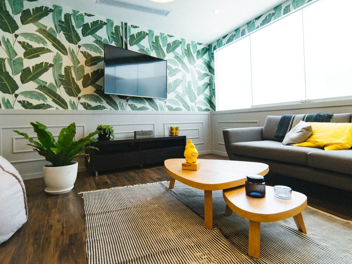 A bright, modern living room with a leafy wallpaper, a wall-mounted TV, a gray sofa with yellow pillows, wooden coffee tables, and a potted plant.