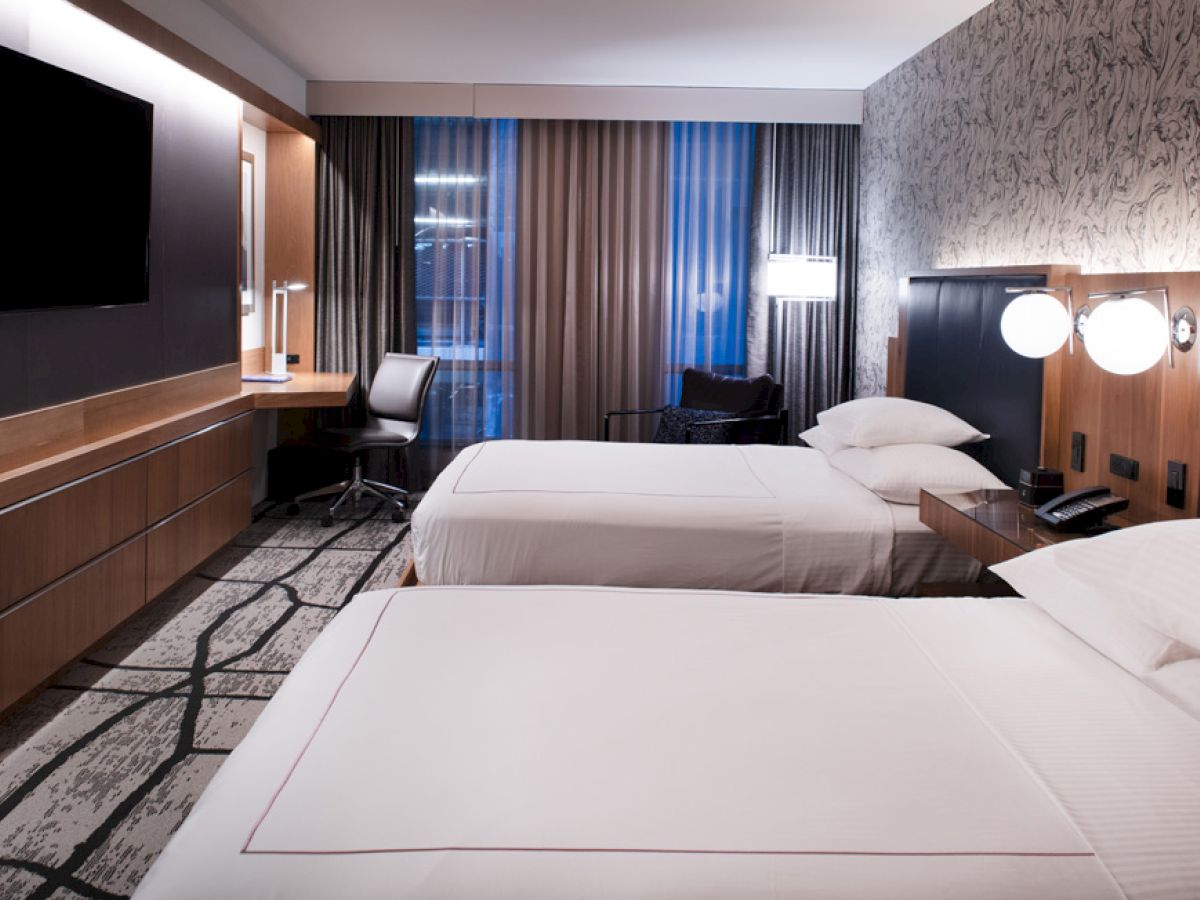 The image shows a modern hotel room with two neatly made double beds, a flat-screen TV, a desk with a chair, and stylish decor.