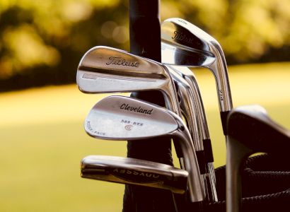 A close-up photo of golf clubs in a bag, with brands such as Titleist and Cleveland clearly visible, set against a blurred outdoor background.