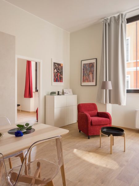The image shows a bright, modern living room with a dining area, kitchen, red armchairs, a red checkered sofa, large window, and wall art.