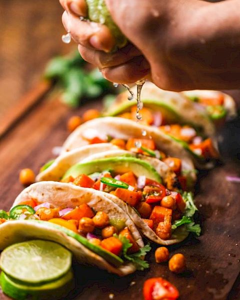 The image shows a hand squeezing lime over a wooden board with several deliciously garnished tacos, featuring ingredients like chickpeas and fresh vegetables.