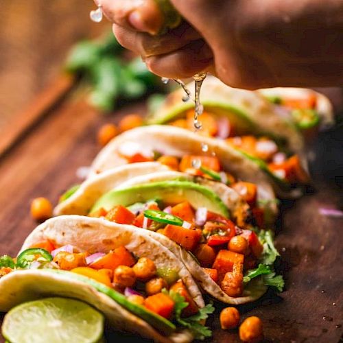 The image shows a hand squeezing lime over a wooden board with several deliciously garnished tacos, featuring ingredients like chickpeas and fresh vegetables.