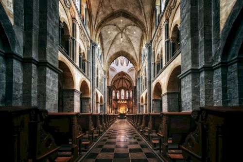 This image depicts the interior of a grand Gothic-style church or cathedral, with high vaulted ceilings, arched columns, and wooden pews along the aisle.