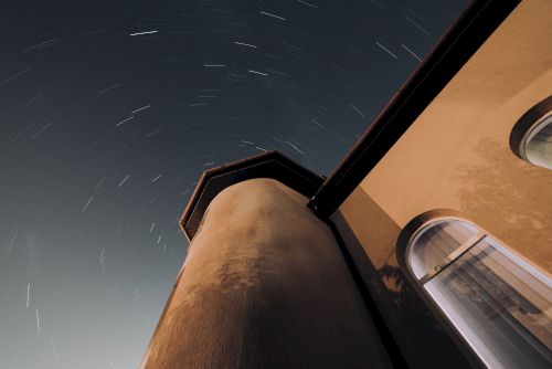 The image shows a building with a tower, possibly a lighthouse, under a night sky with star trails indicating long-exposure photography.