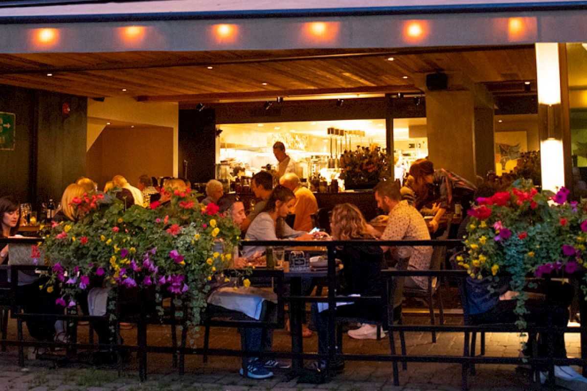 Outdoor restaurant seating area at night with several people dining. The area is decorated with colorful flower arrangements.