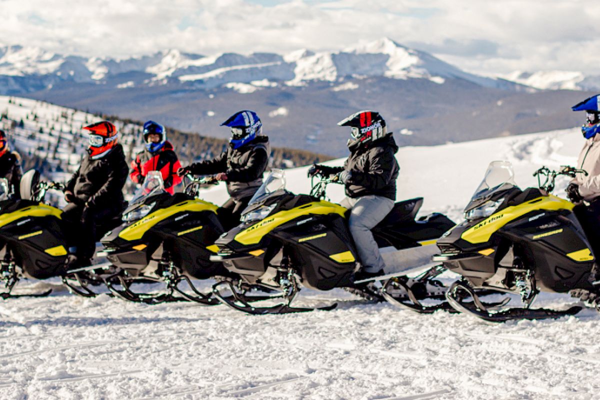A group of people on snowmobiles are receiving instructions from a person in a red jacket, set against a snowy mountainous background.