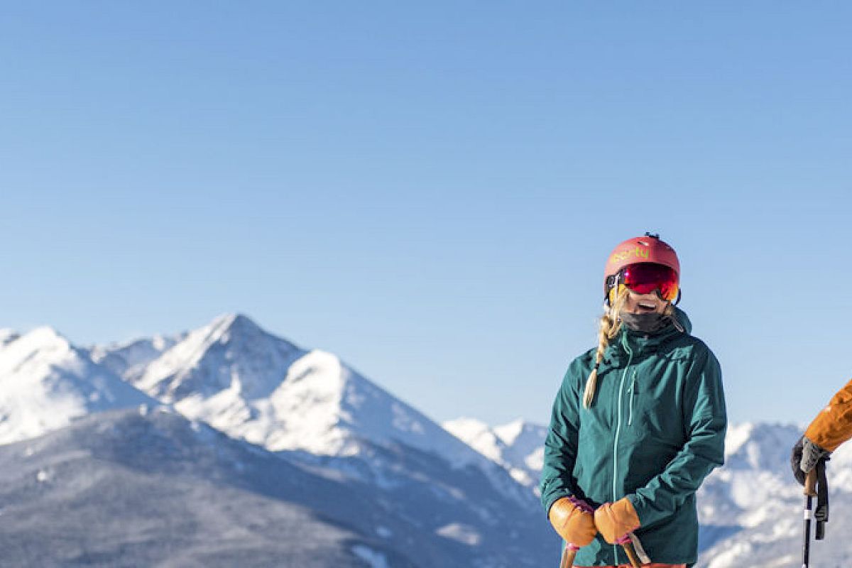 Two people in ski gear stand on a snowy mountain with scenic snow-covered peaks in the background, under a clear blue sky.