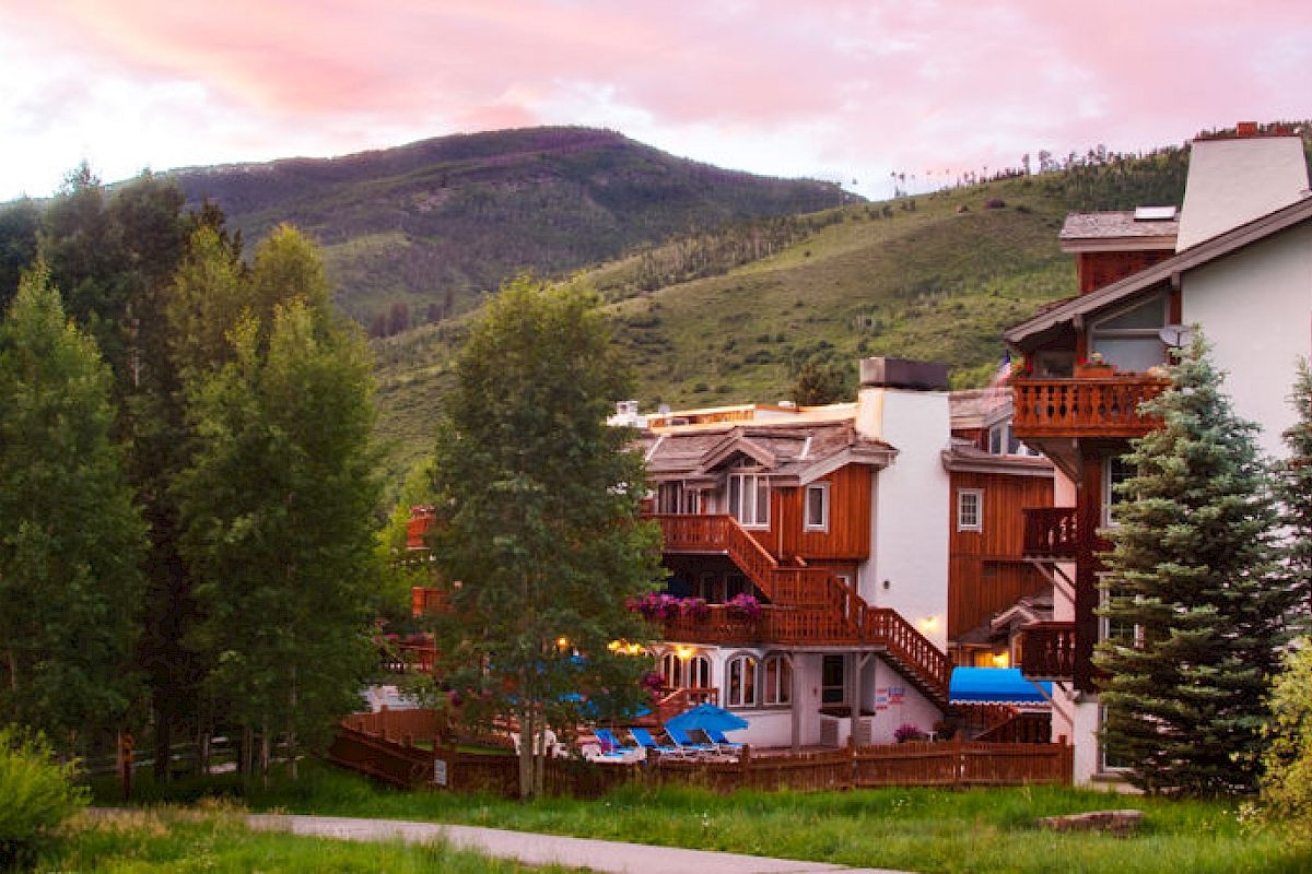 A picturesque scene featuring a rustic building surrounded by lush trees and greenery, with hills in the background under a colorful sky.