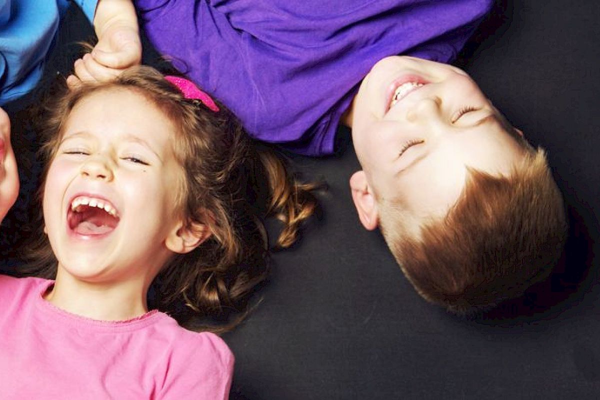 Three children in colorful clothes lie on their backs on a dark surface, laughing and holding hands, sharing a joyful moment together.
