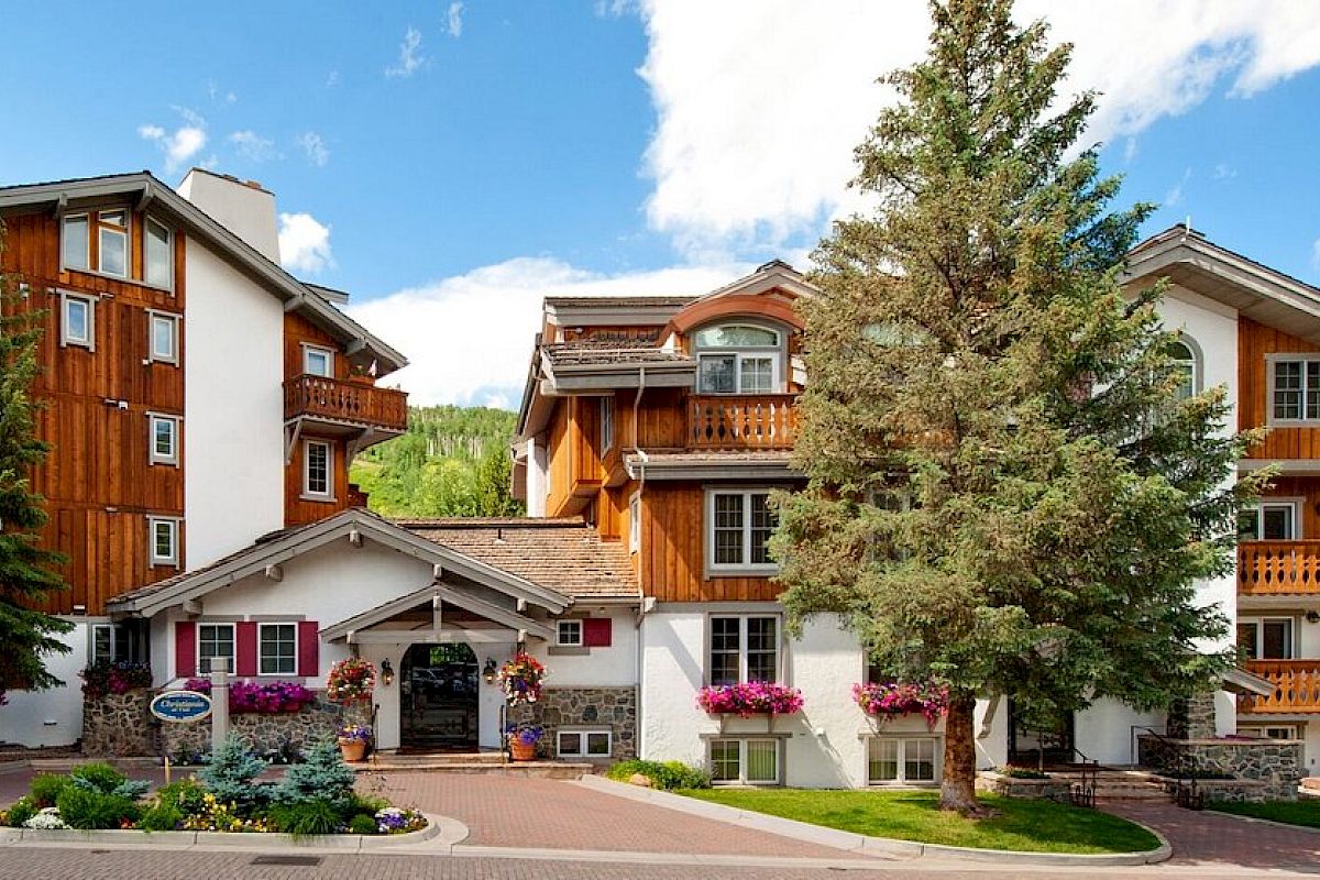 The image shows a picturesque building with wood and white facades, surrounded by trees and colorful flowers, under a bright blue sky.