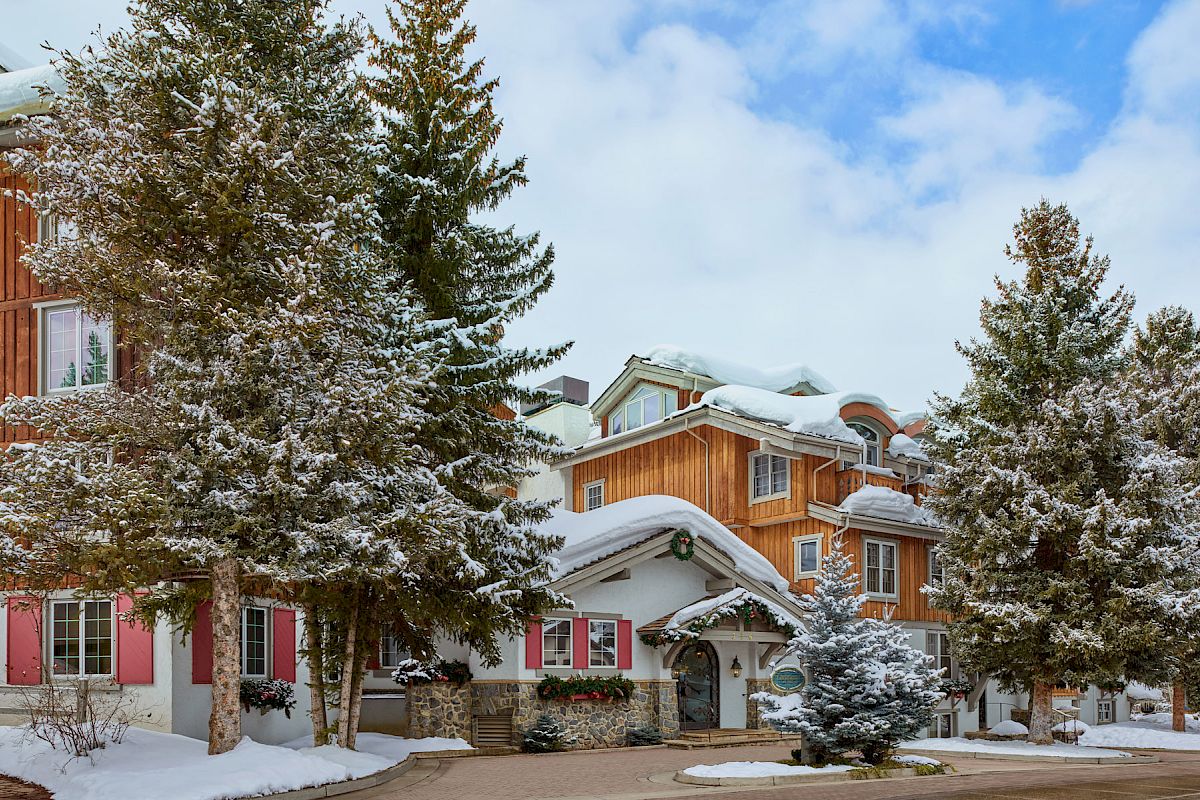 A picturesque, snow-covered wooden lodge with pink shuttered windows, surrounded by evergreen trees and a clear blue sky.