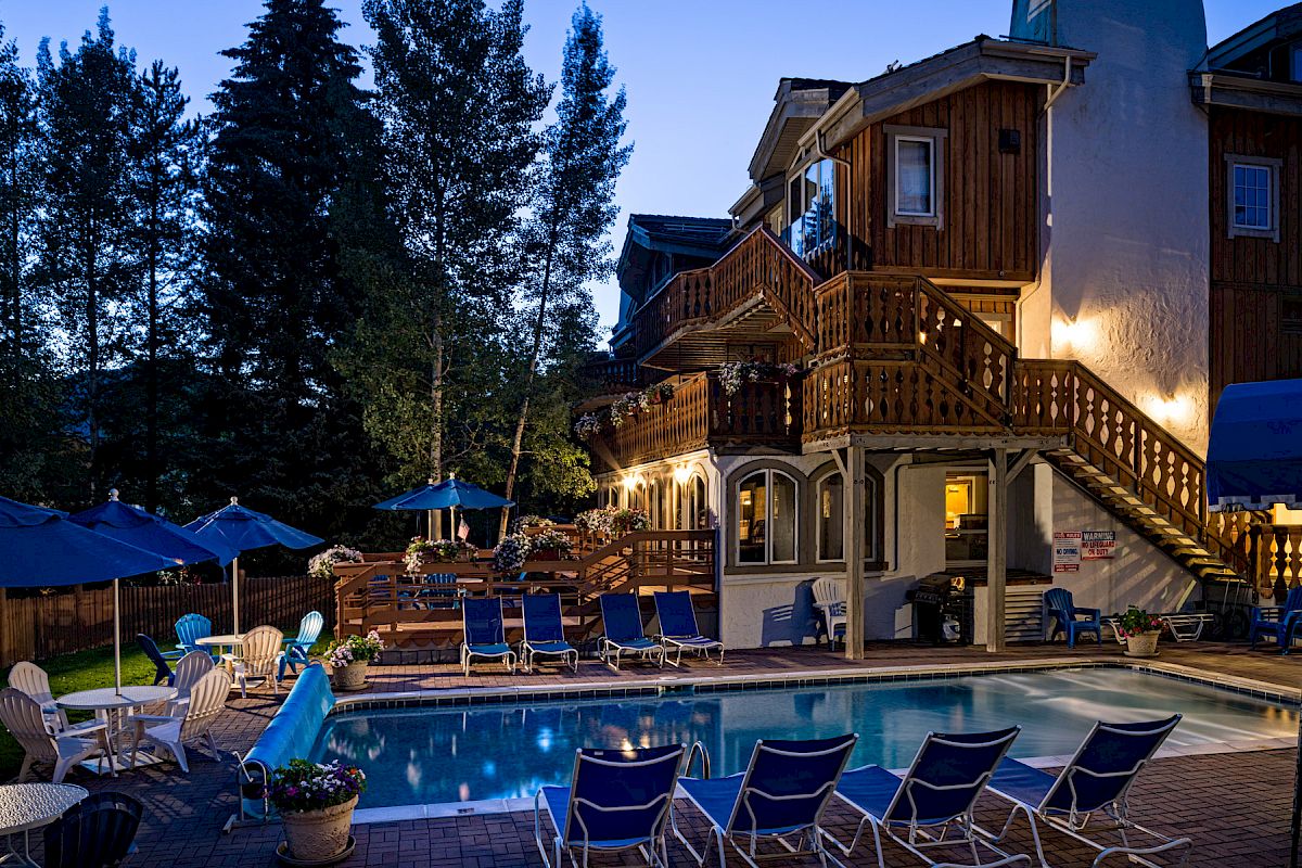 An evening view of a quaint hotel with lit windows, a pool surrounded by lounge chairs, and umbrellas, nestled among tall trees.