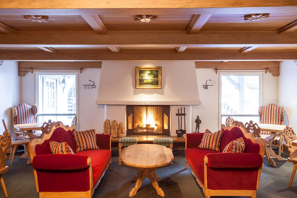 A cozy room with red-cushioned wooden furniture, a central coffee table, and a lit fireplace, flanked by two windows and chairs.