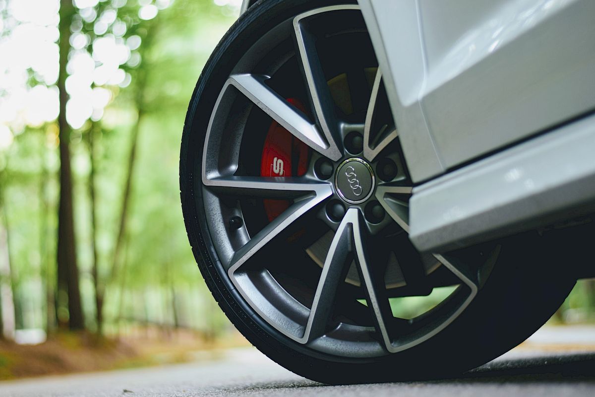 The image shows a close-up of a car wheel with a stylish alloy rim and red brake caliper, outdoors with blurred trees in the background.