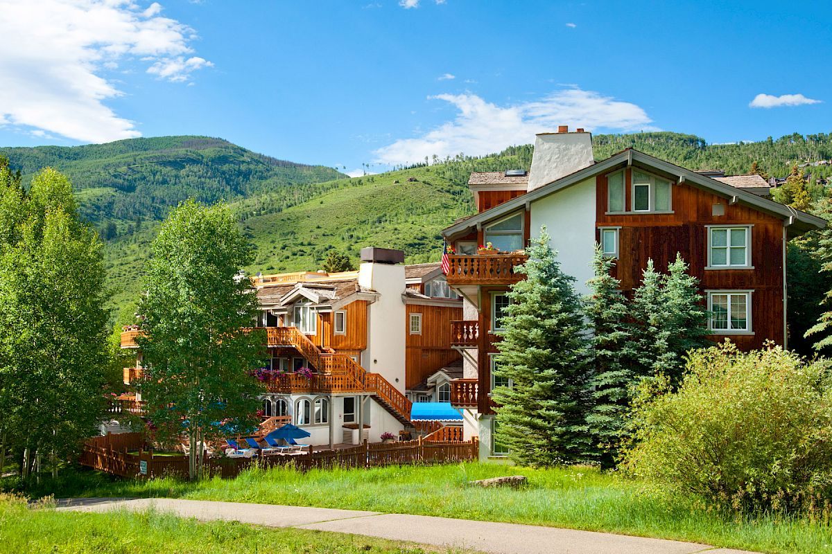 A picturesque mountain lodge with wooden balconies stands amid lush greenery and trees, surrounded by scenic hills and a clear blue sky.