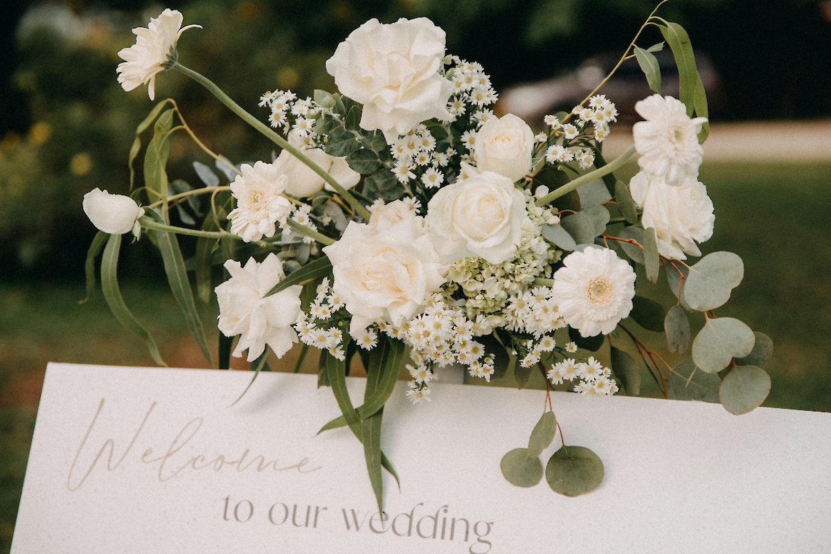A white floral wedding bouquet with roses and baby's breath sits atop a &ldquo;Welcome to our wedding&rdquo; sign, creating a romantic welcome.