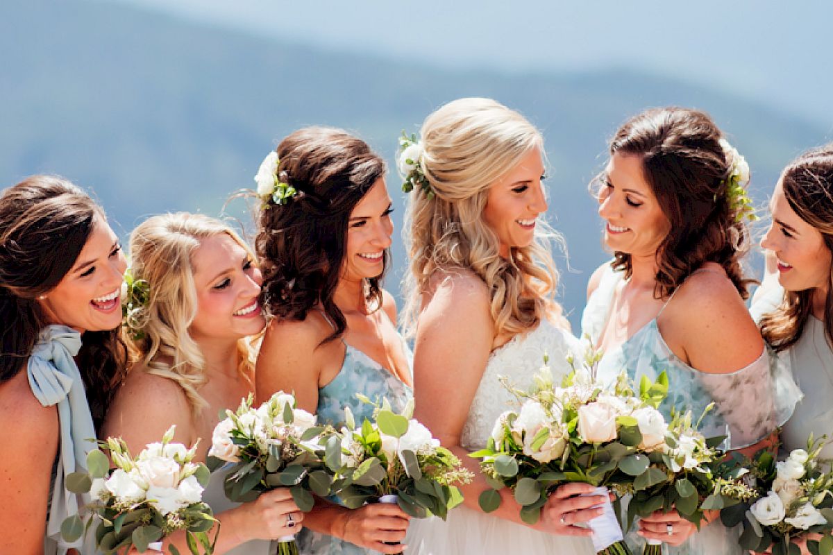 A group of six women, likely a bride and her bridesmaids, standing close together holding bouquets, smiling, and dressed in light-colored dresses.