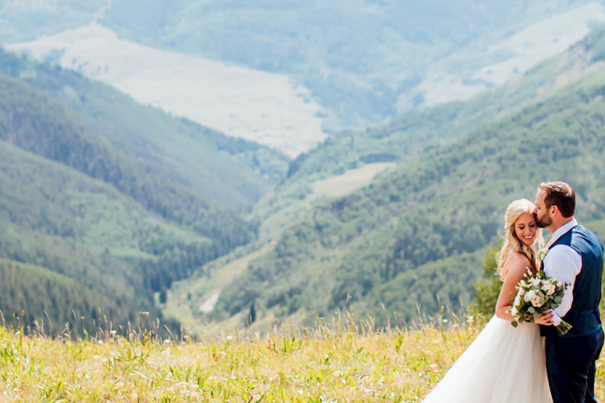 A bride and groom embrace in a beautiful mountainous landscape, with lush green hills and a distant valley forming the backdrop.