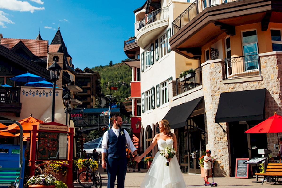 A couple in wedding attire walks hand in hand through a lively outdoor plaza with colorful umbrellas and charming buildings.