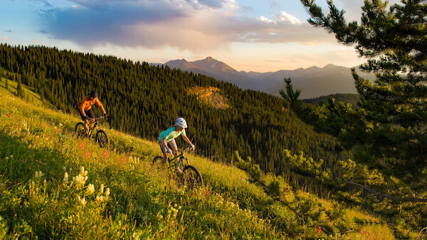 Two people are mountain biking on a lush, green hillside with a scenic forest and mountains in the background under a partly cloudy sky.
