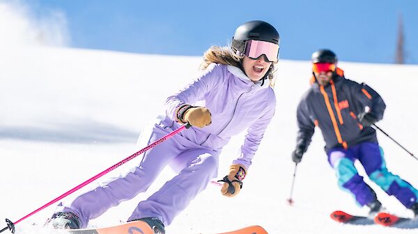Two skiers carve down a snowy slope, one in a lavender suit leading, the other in dark gear following close behind, against a bright blue sky.