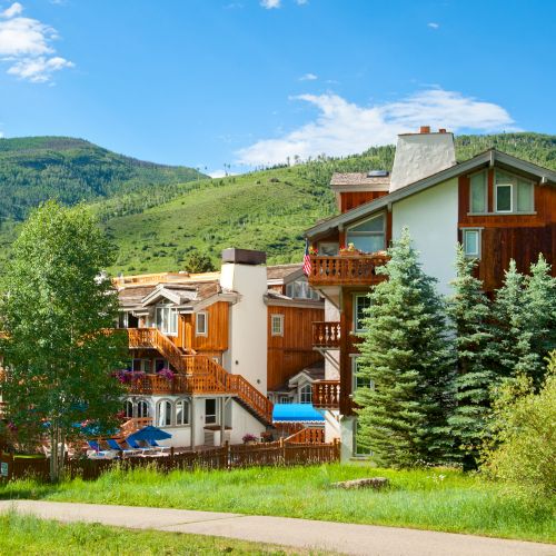 A scenic view of a wooden lodge surrounded by lush greenery, trees, and mountains under a clear blue sky on a sunny day.