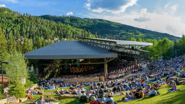An outdoor concert venue is filled with an audience enjoying a performance, set against a backdrop of lush green hills and a partly cloudy blue sky.