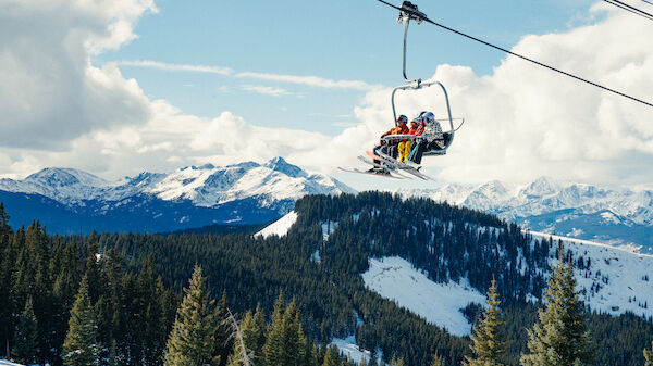 A ski lift carries three skiers over snow-covered mountains and pine trees under a bright blue sky with clouds.
