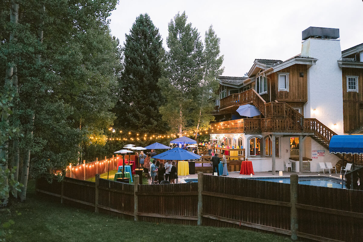The image shows a backyard gathering with string lights, umbrellas, and people enjoying a poolside atmosphere near a house.