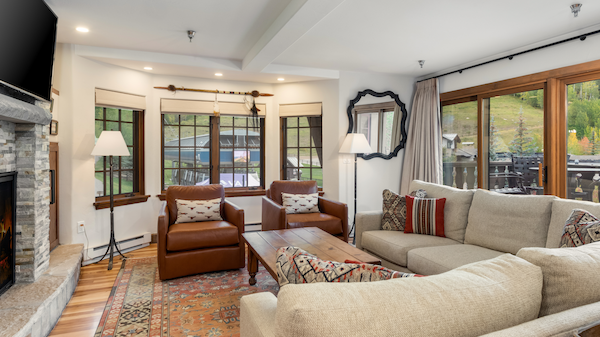 A cozy living room with a beige sectional, brown leather chairs, a patterned rug, large windows, a stone fireplace, and rustic decor.