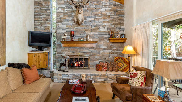 Cozy living room with a stone fireplace, mounted deer head, wooden shelves, and elegant furnishings, featuring warm, earthy tones.