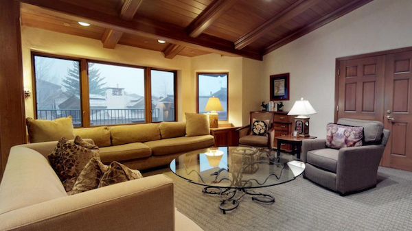 The image shows a cozy living room with a large sofa, glass coffee table, and windows providing natural light. It has a warm atmosphere.