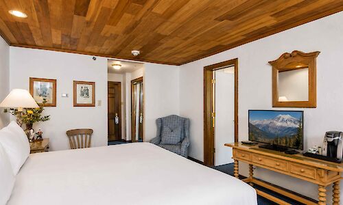 A cozy hotel room with a large bed, wooden ceiling, framed art, a small desk with a TV, a coffee maker, and a blue armchair in the corner.