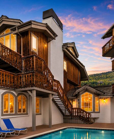 A charming hotel with wooden balconies, arched windows, and a small pool surrounded by lounge chairs; sunset paints the sky in pink and orange.