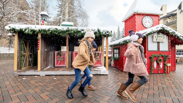 People in winter clothing walk past festive, snow-covered buildings with Christmas decorations and a clock tower in a snowy setting.