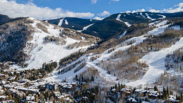A snow-covered mountain landscape with ski slopes and a town below, framed by trees and a blue sky with fluffy clouds.