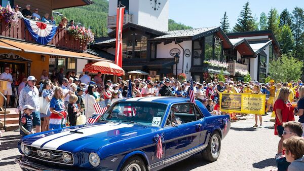A blue classic car drives through a cheering crowd at a sunny outdoor event near a quaint village square, with onlookers and banners.