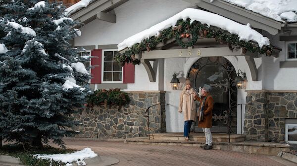 A couple stands outside a snowy home, adorned with holiday decorations, enjoying a moment together. It looks cozy and festive.