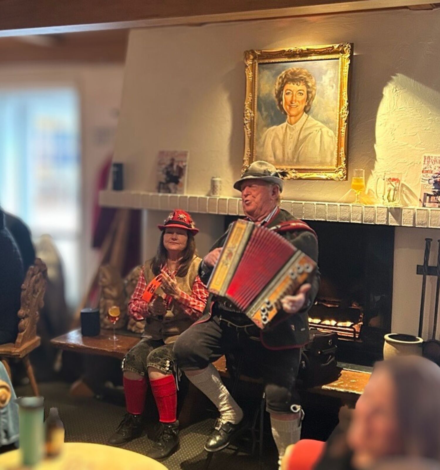 A cozy cafe scene with a man playing an accordion as two people in festive outfits sit nearby, while a portrait hangs above a lit fireplace.