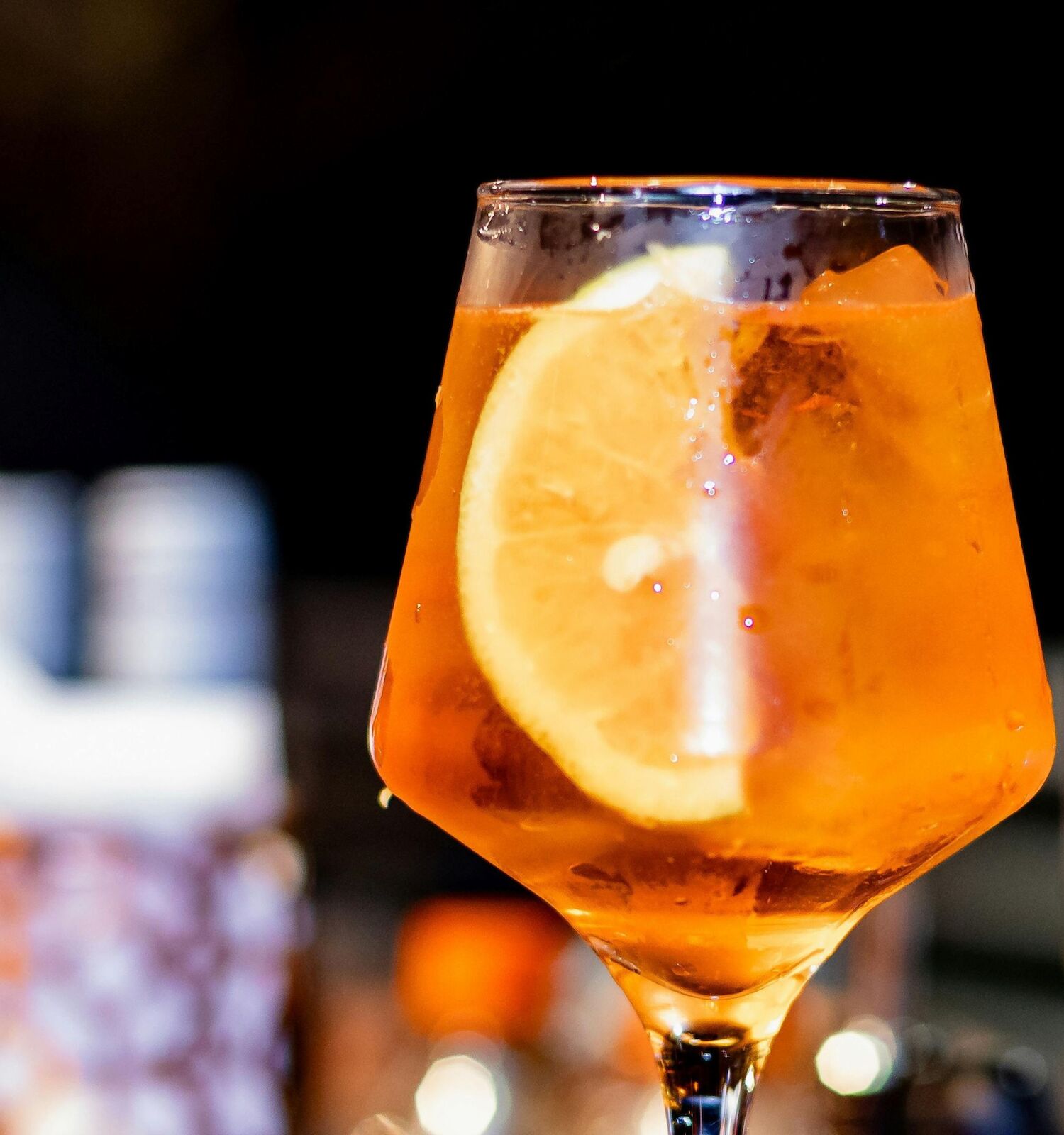 An orange cocktail in a stemmed glass with a large orange slice, ice, and a blurred bar background.