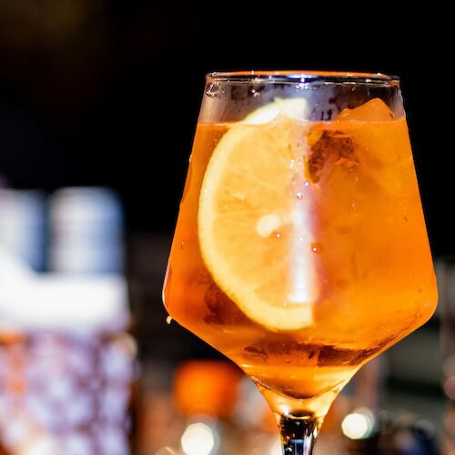 A stylish orange cocktail with a slice of orange garnish in a stemmed glass, vibrant against a dark bar backdrop.