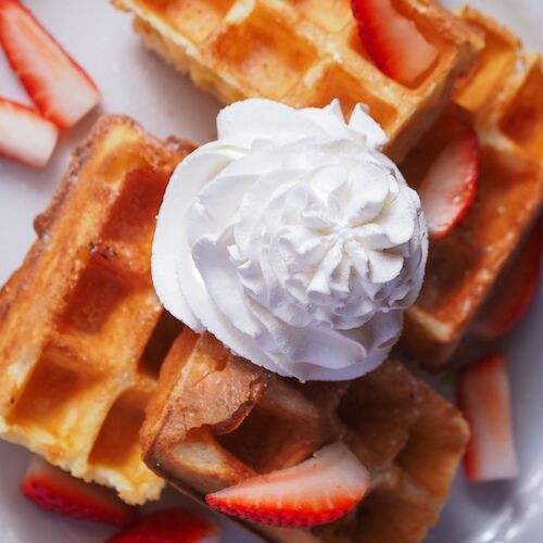 Waffles topped with sliced strawberries and a dollop of whipped cream, drizzled with syrup, on a white plate.