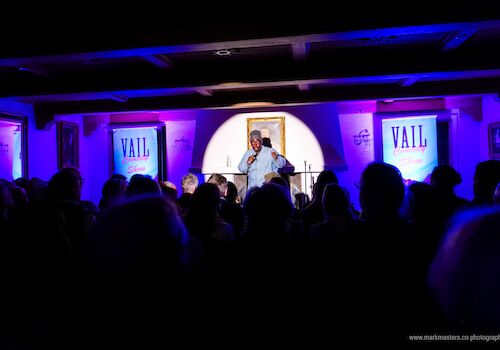 A man performs on a stage in a dim, purple-lit venue with a crowd watching; banners say "VAIL" in the background, framed by a bright central spotlight.