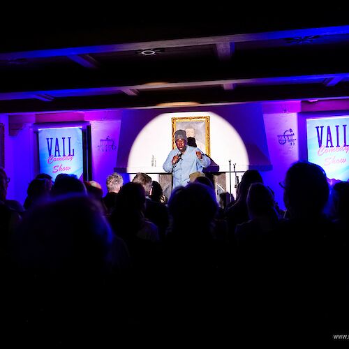 A man performs on a stage in a dim, purple-lit venue with a crowd watching; banners say "VAIL" in the background, framed by a bright central spotlight.