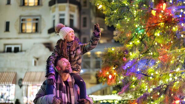 A joyful family couple with a child on shoulders admires a brightly lit Christmas tree in a festive city square at night.