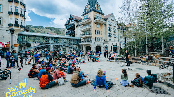 A lively outdoor comedy show in a European-style town square, with a crowd seated on the pavement, a clown on a stage, and festival branding in the corner.