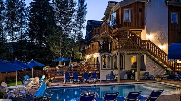 A resort-style hotel with a wooden, multi-story building and a swimming pool surrounded by lounge chairs at dusk, with trees in the background.